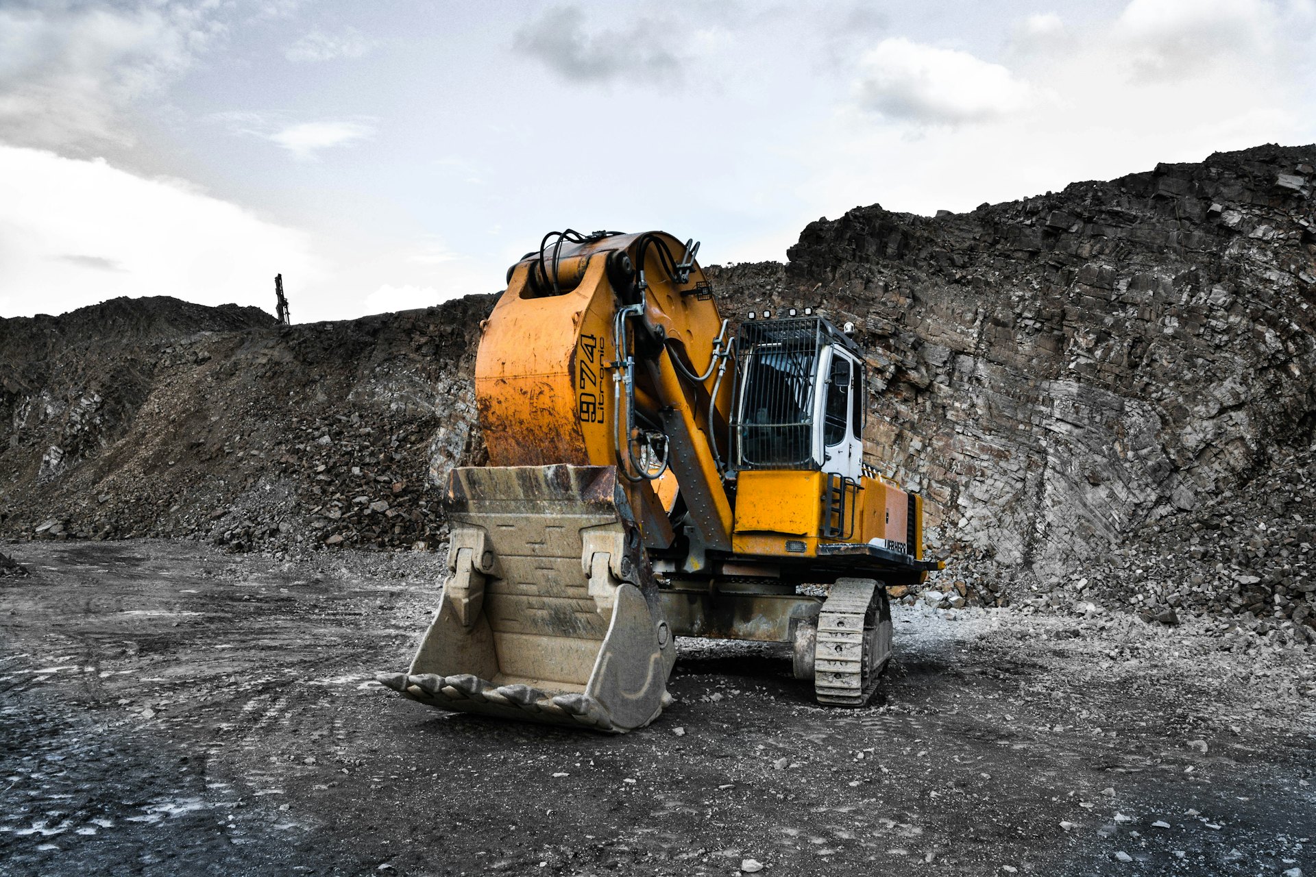 An excavator standing in a working stone quarry, dark rock face behind, gravel underfoot.